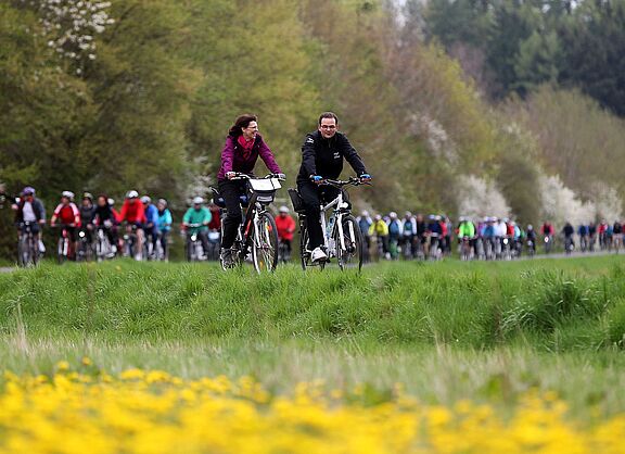 Zu sehen ist eine Gruppe Radfahrern, im Vordergrund Blumenwiese und im Hintergrund Bäume