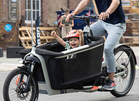 Ein Kind in einem Lastenrad hebt jubelnd die Arme, das Lastenrad fährt eine erwachsene Person in Jeans. für den Kindertransport ist der Traum vieler junger Familien.